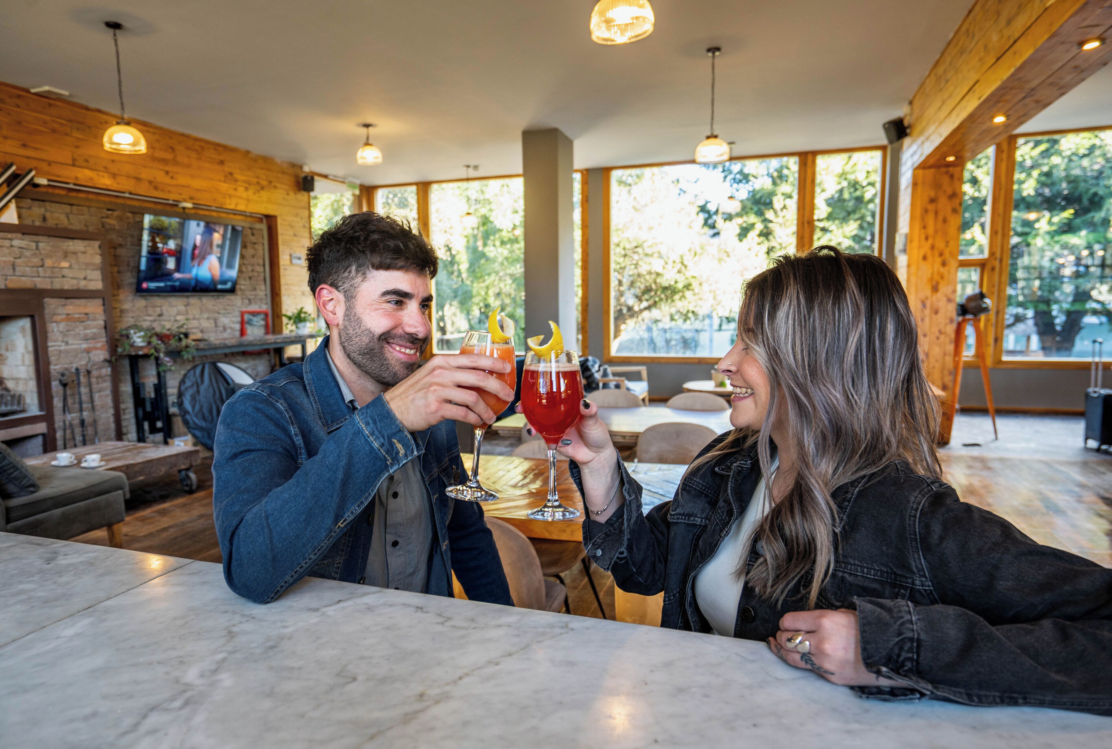 Un couple trinque avec des boissons à une table dans une pièce moderne et bien éclairée, dotée de grandes fenêtres et d’un téléviseur en arrière-plan.
