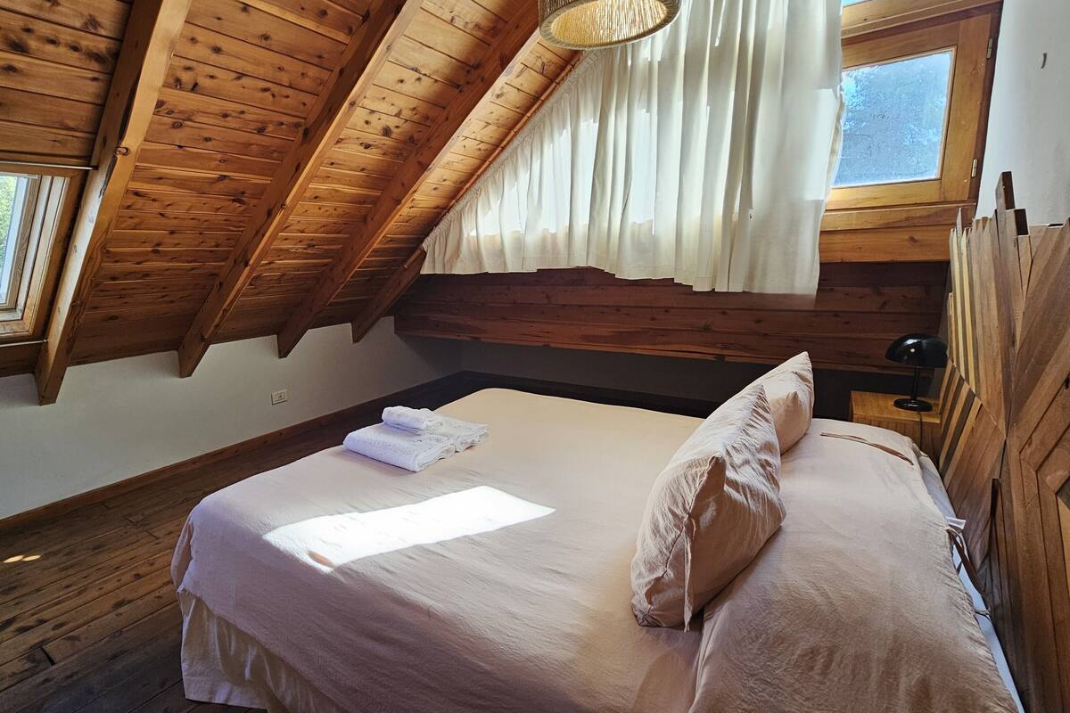 Ein Schlafzimmer mit Holzboden, einem Bett mit Kissen, einem Fenster mit Vorhängen und einer Nachttischlampe.