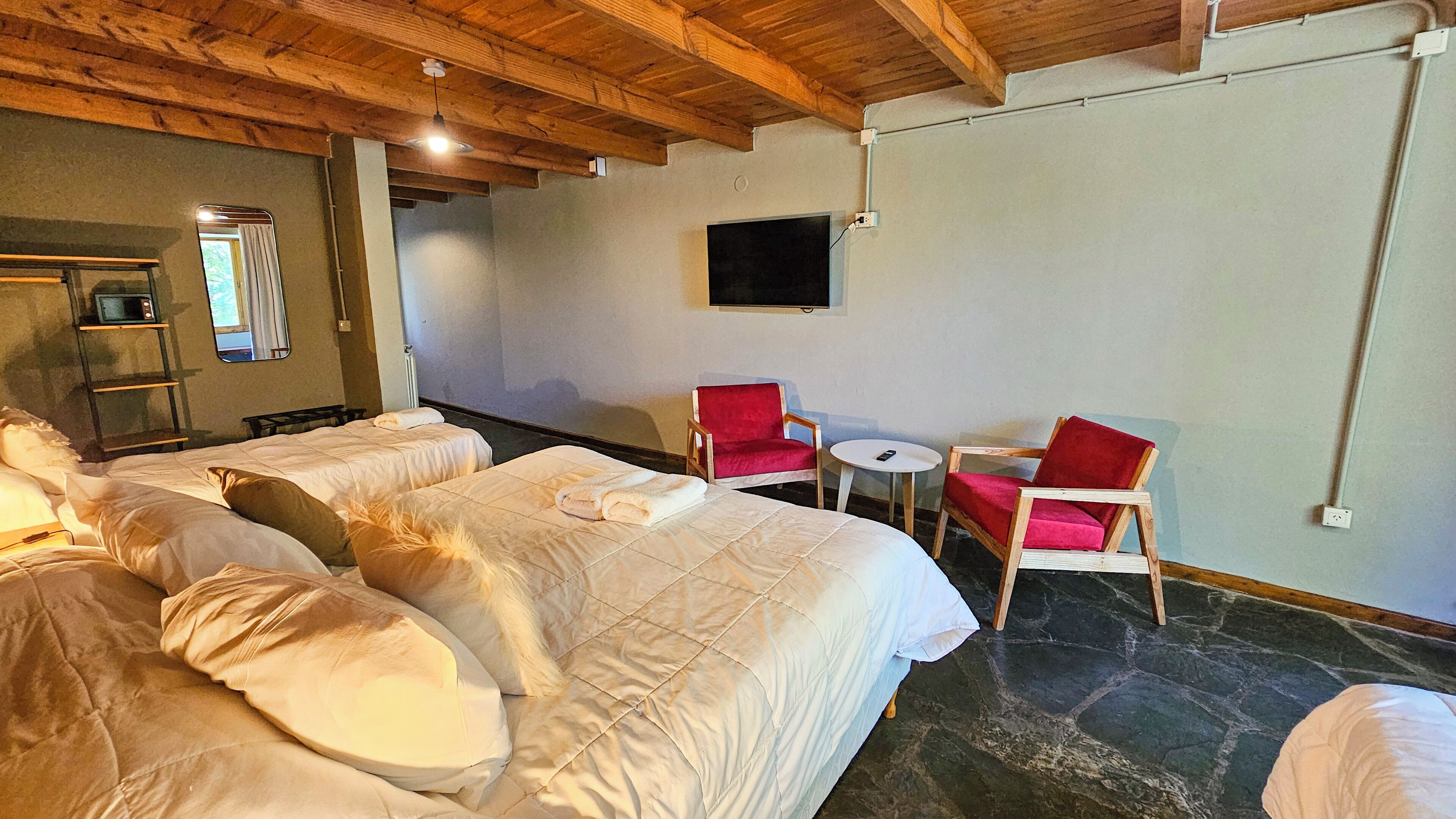 A hotel room with two beds, a small table, two red chairs, a TV on the wall, and a wooden ceiling.