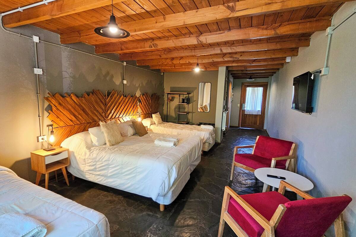 Une chambre d’hôtel avec un plafond en bois, un grand lit, deux petits lits, une chaise rouge, une petite table et un miroir au mur.