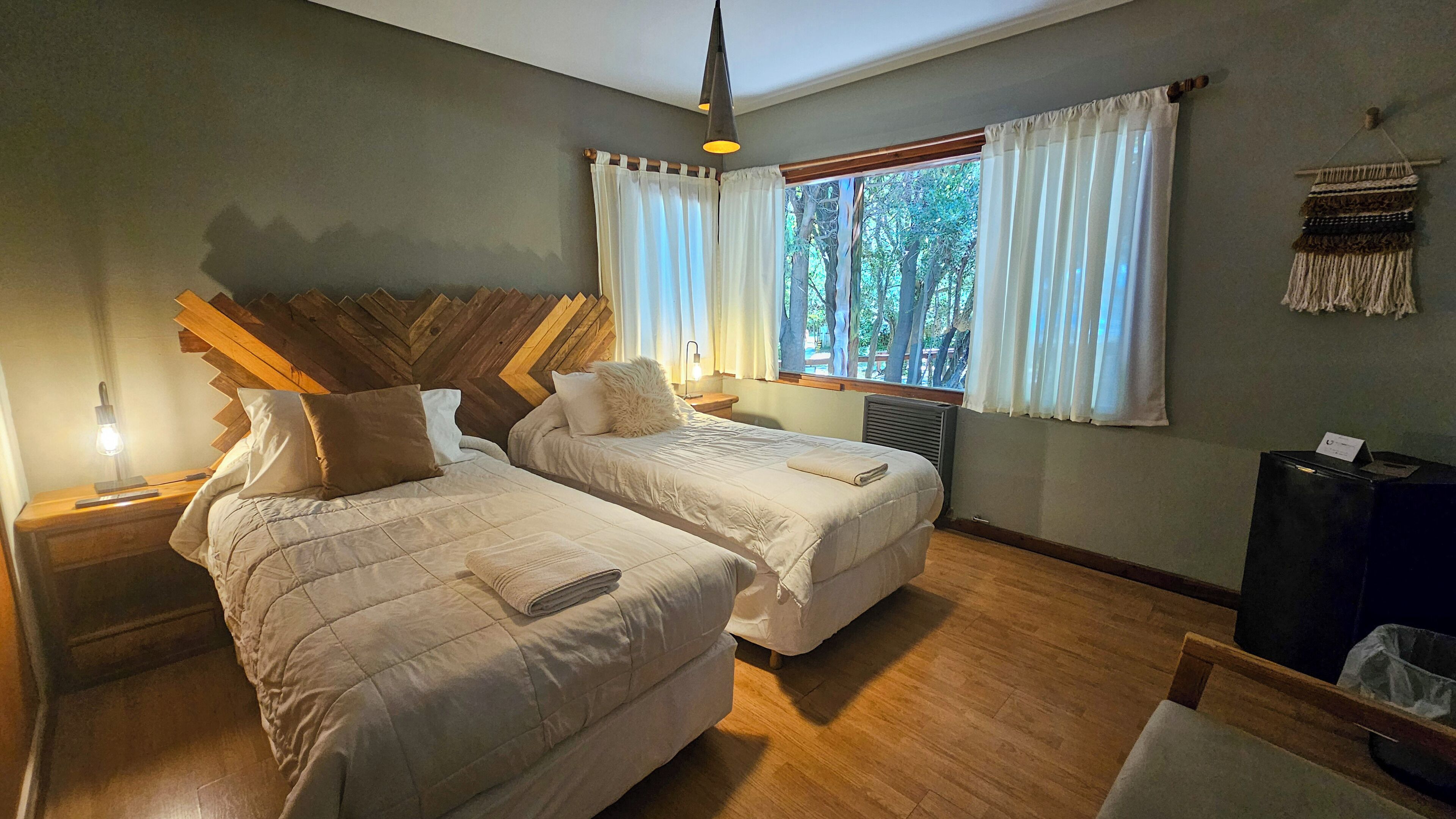 A bedroom with two beds, a wooden headboard, a nightstand with a lamp, a window with curtains, and a wall-mounted tapestry.