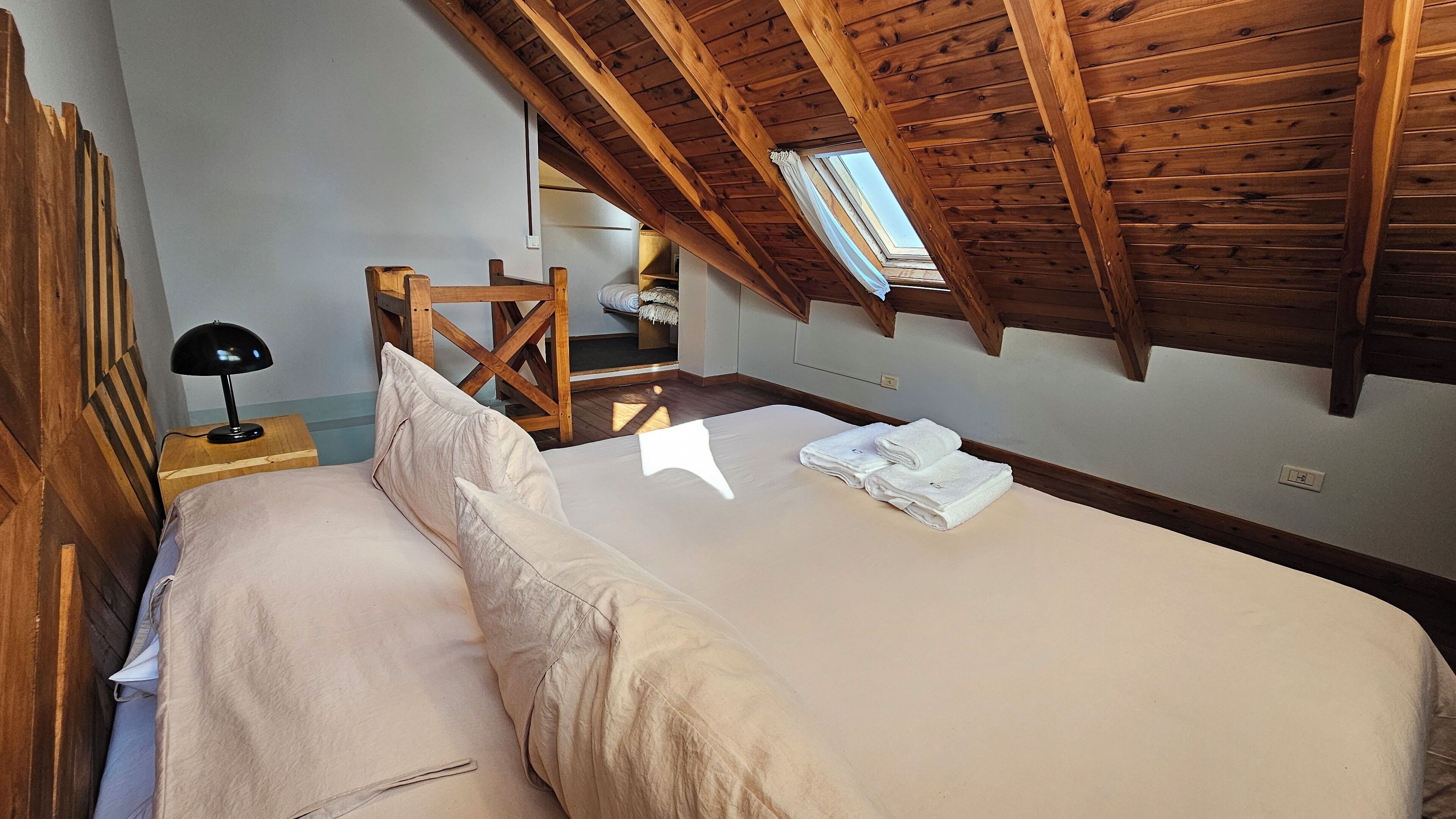A bedroom with a wooden ceiling, a bed with white linens, a nightstand with a lamp, and a window with curtains.