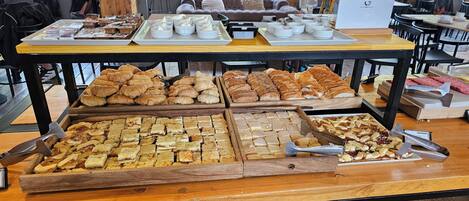 Una mesa de buffet con diversos pasteles y postres, incluyendo croissants y tartas, en un ambiente de cafetería.