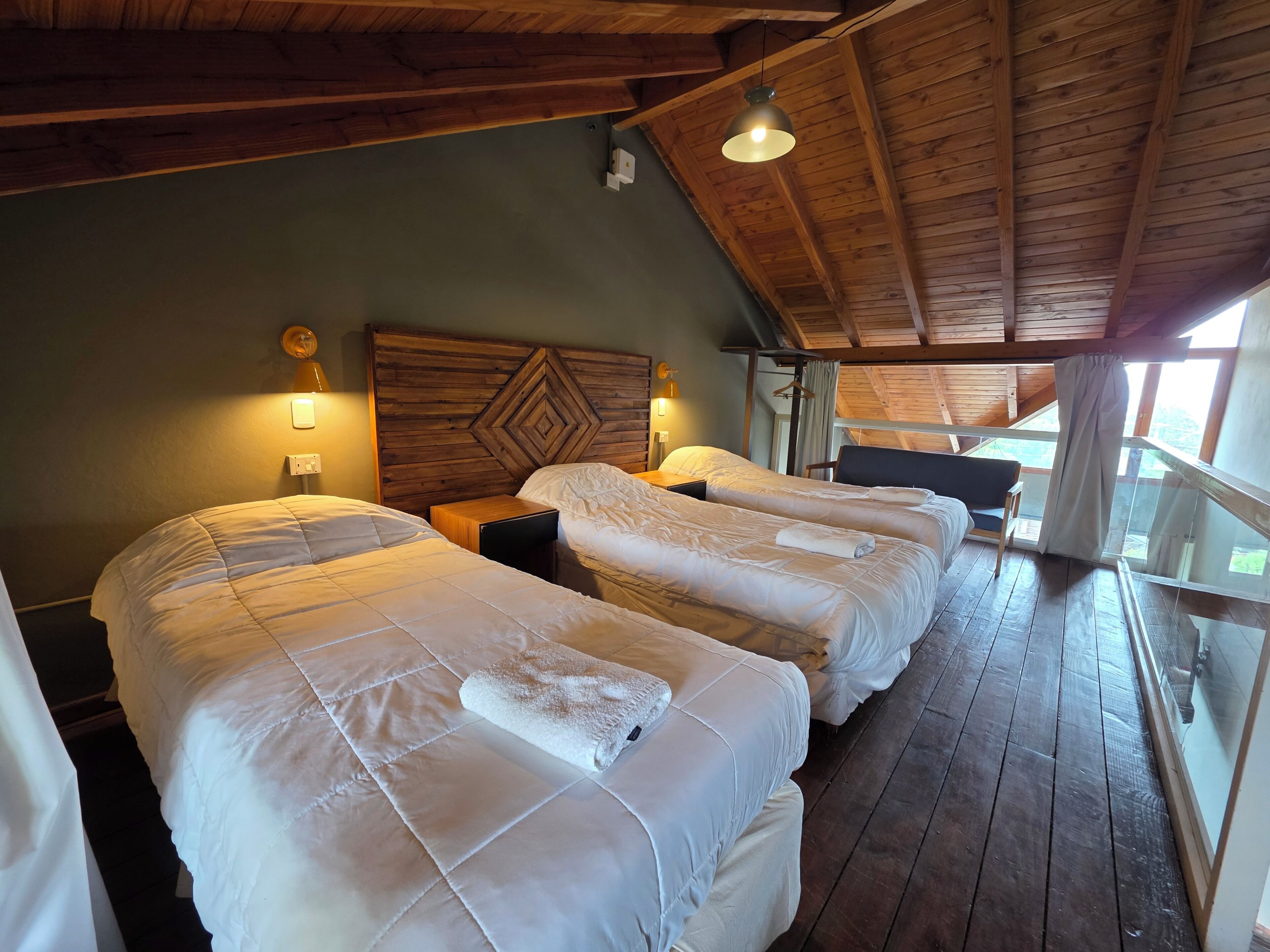 A room with three beds, wooden ceiling beams, a hanging light fixture, and a view through a glass railing.