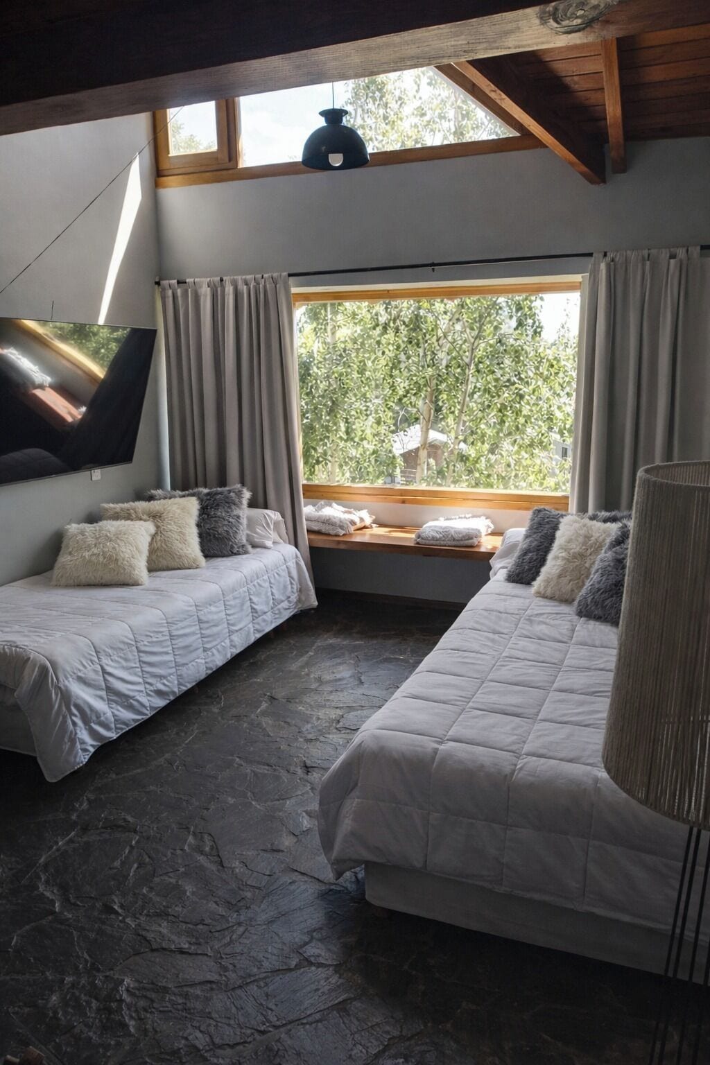 A room with two beds, a large window with curtains, a flat-screen TV, and a stone floor.