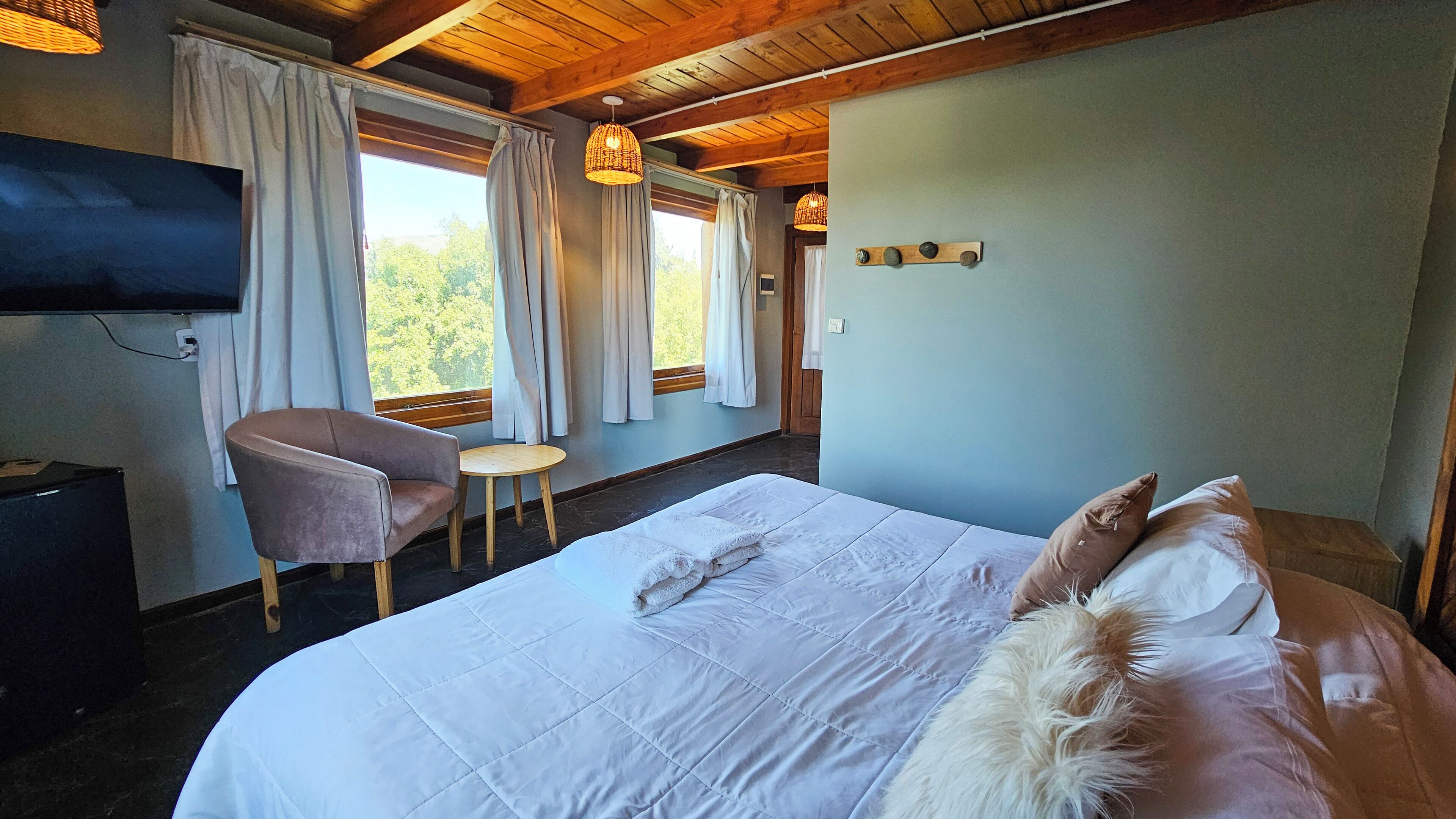 Un dormitorio con cama, una silla, una mesita, televisor y ventanas con cortinas.