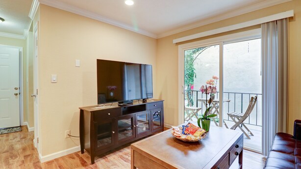 Una sala de estar con una mesa de centro de madera, un televisor de pantalla plana sobre un mueble bajo y un balcón con una silla y una planta en maceta.