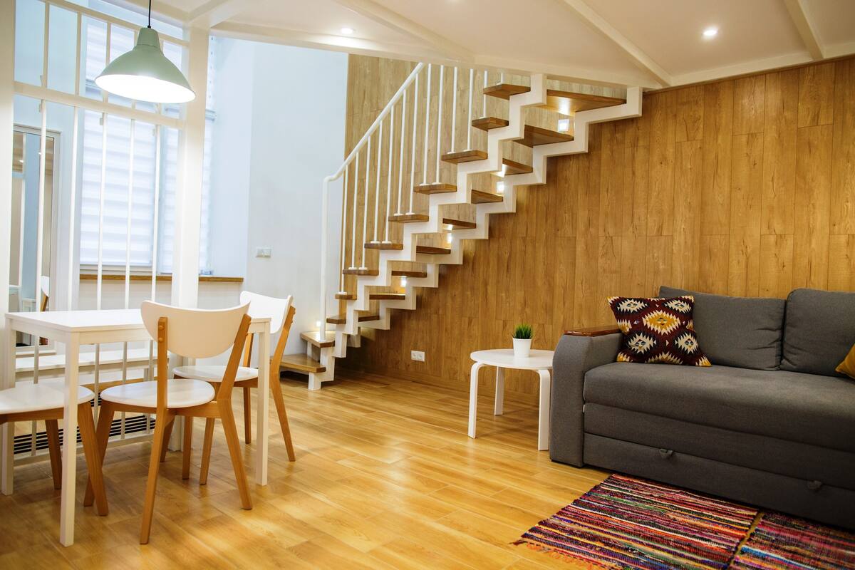A modern living room with a wooden staircase, a gray sofa, a white dining table with chairs, and a colorful rug.