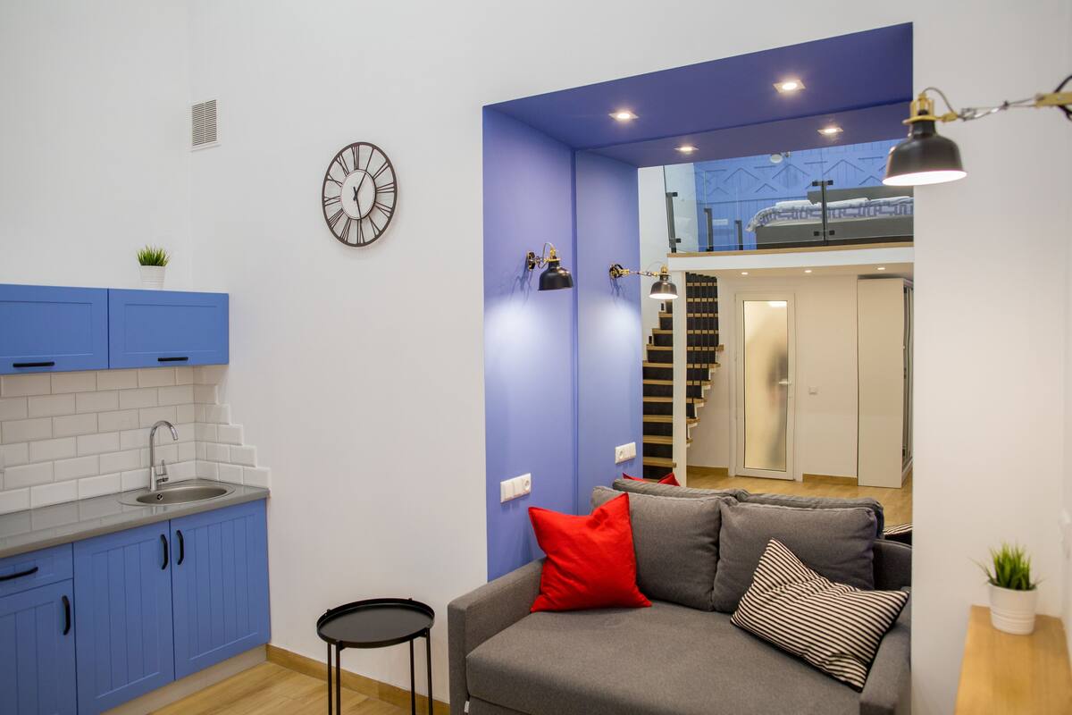 A compact living space with a kitchenette, a grey sofa with red and striped pillows, a small round table, and a wall-mounted clock.