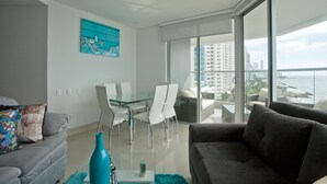 A modern living room with a glass dining table, white chairs, a grey sofa, and a large window offering a city view. - Murano Elite Luxury Bocagrande (Cartagena)