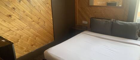 A room with a wooden sloped ceiling, a bed with white bedding, a bedside table, and a mirror on the wall.