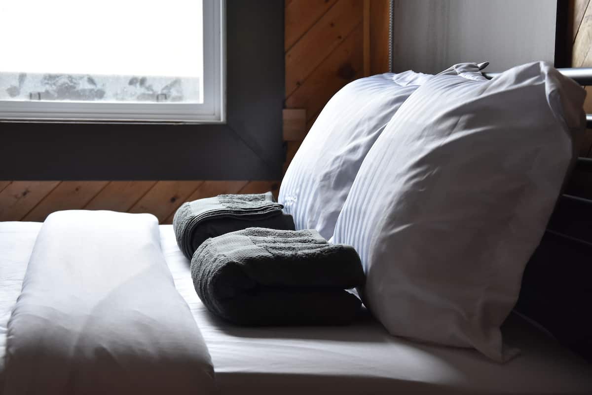 A neatly made bed with white and gray linens, a window with a view, and wooden paneling on the wall.