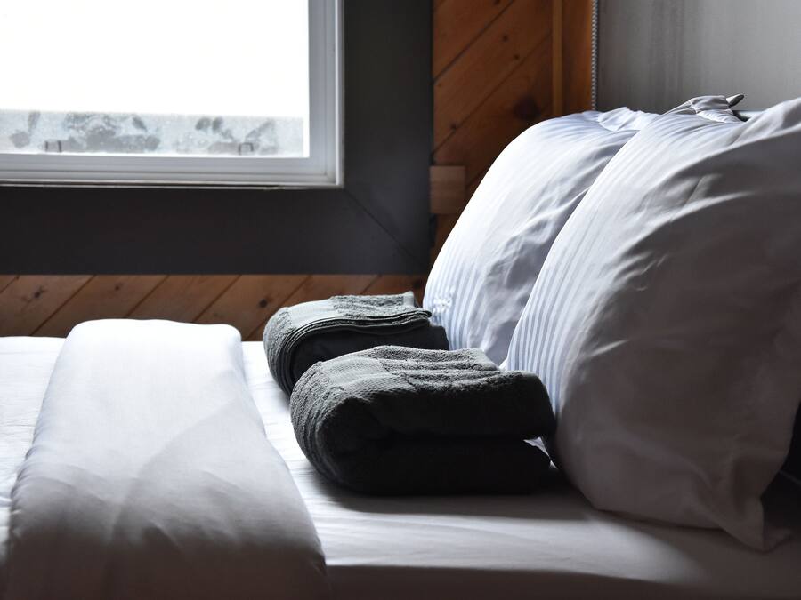 A neatly made bed with white and gray linens, a window with a view, and wooden paneling on the wall.