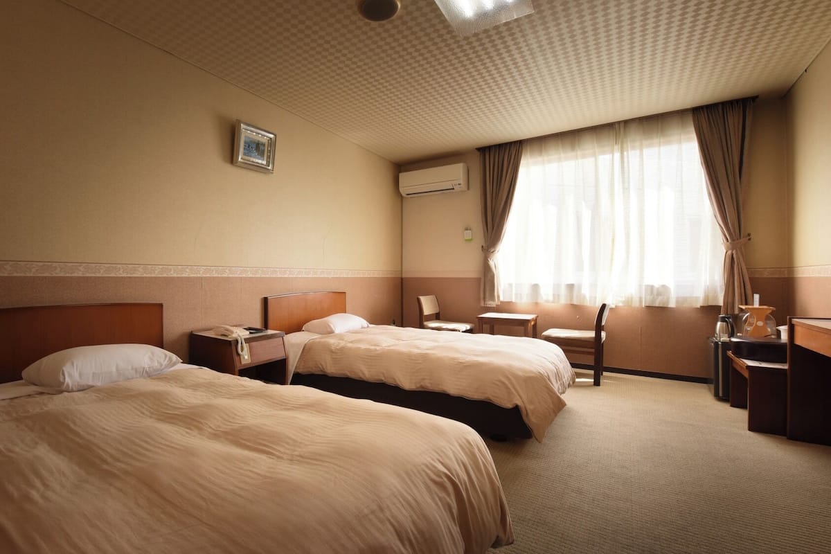 Hotellihuone, jossa on kaksi sänkyä, työpöytä, tuoli, pieni pöytä ja ikkuna, jossa on verhot.