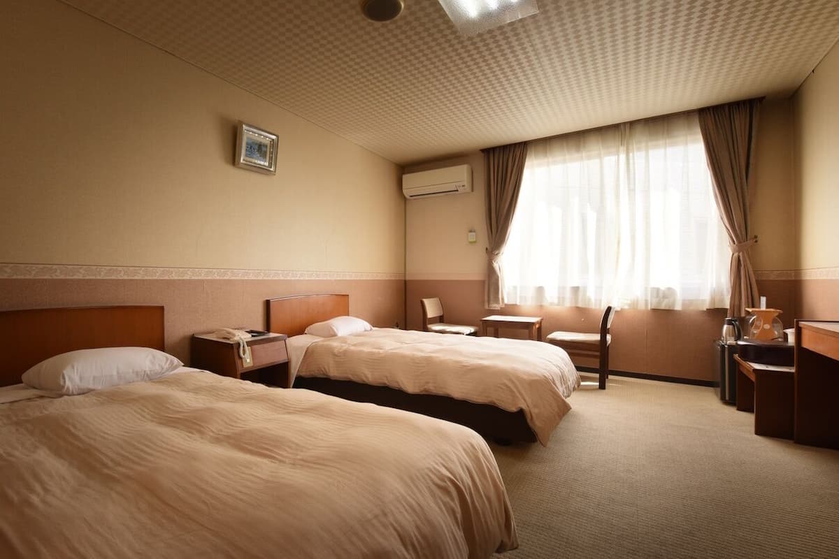 Hotellihuone, jossa on kaksi sänkyä, työpöytä, tuoli, pieni pöytä ja ikkuna, jossa on verhot.