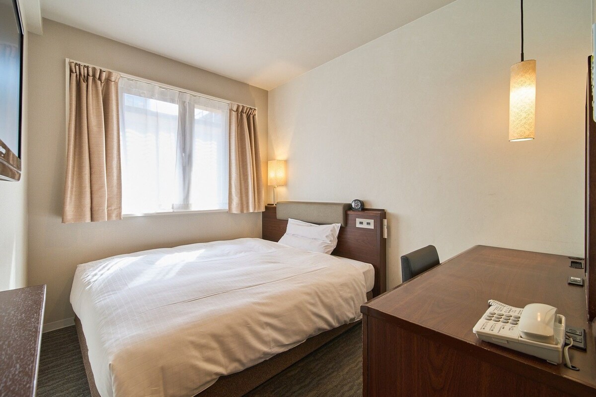 A hotel room with a bed, a desk with a telephone, a window with curtains, and a wall-mounted lamp.