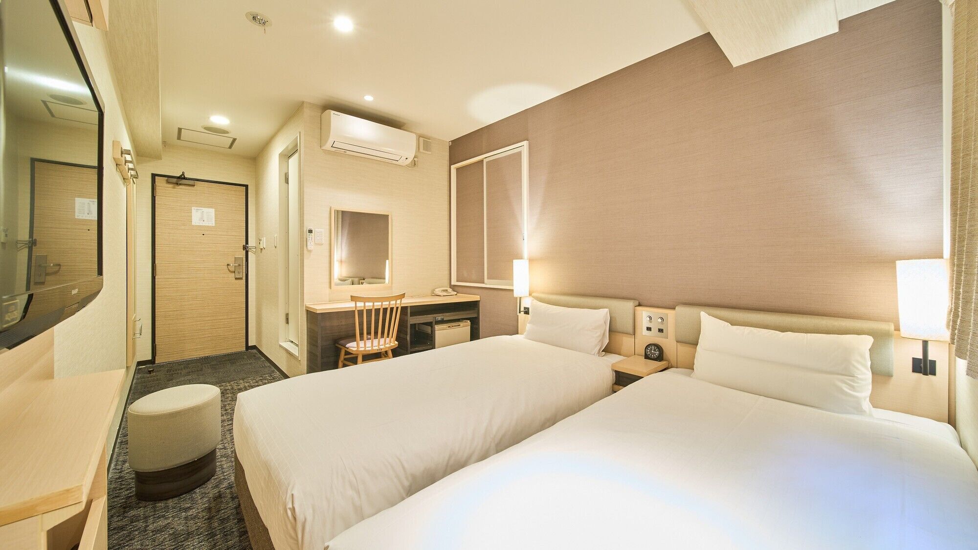 Habitación de hotel con dos camas, un escritorio, una silla, un espejo, tocador y la puerta del baño.