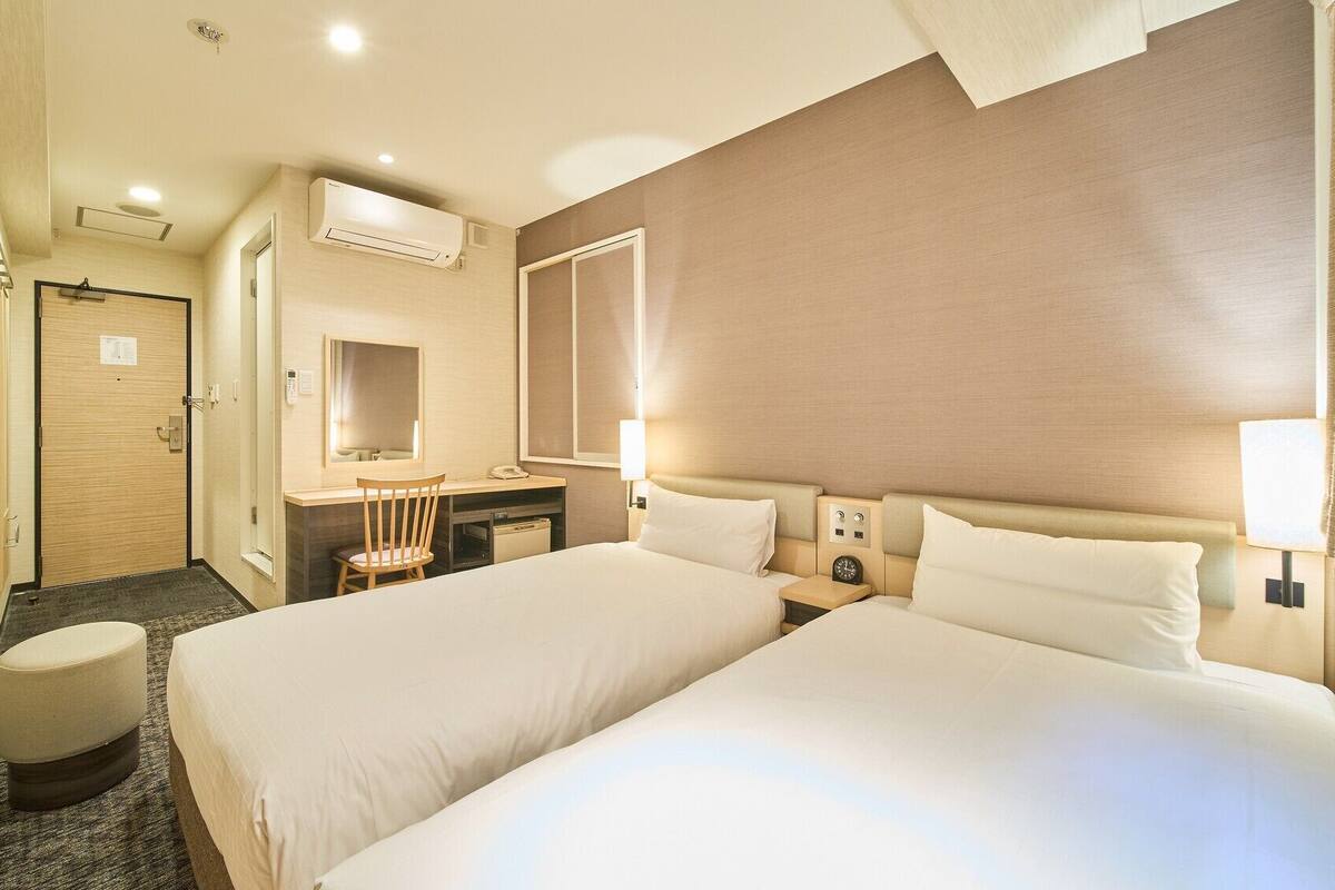 A hotel room with two beds, a desk, a chair, a mirror, a vanity, and a bathroom door.