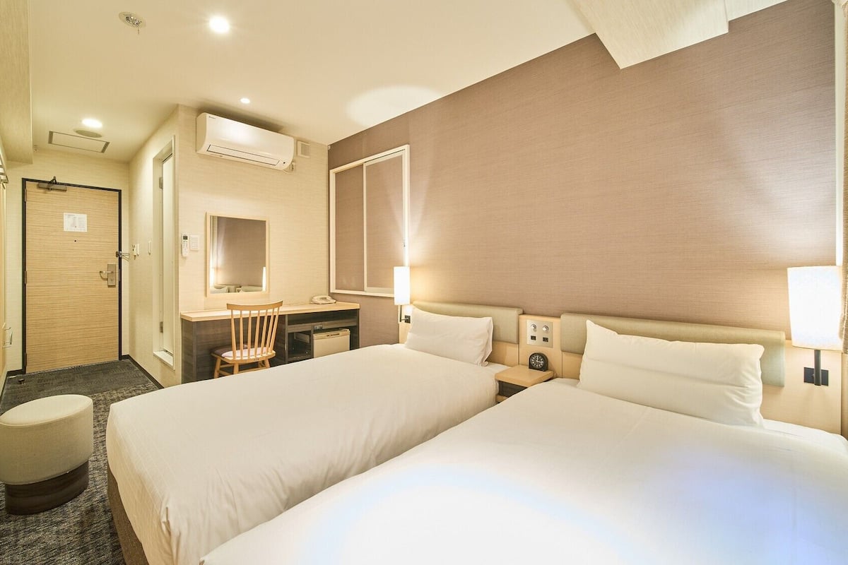 Habitación de hotel con dos camas, un escritorio, una silla, un espejo, tocador y la puerta del baño.