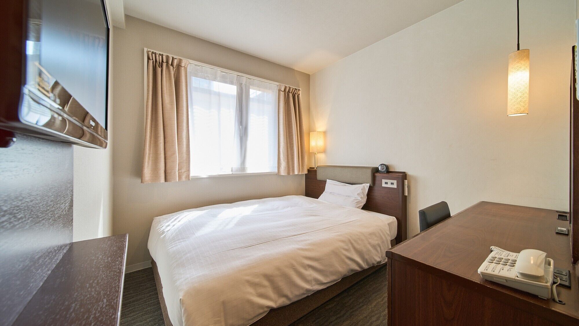 Habitación de hotel con cama, escritorio con teléfono, ventana con cortinas y lámpara fijada a la pared.