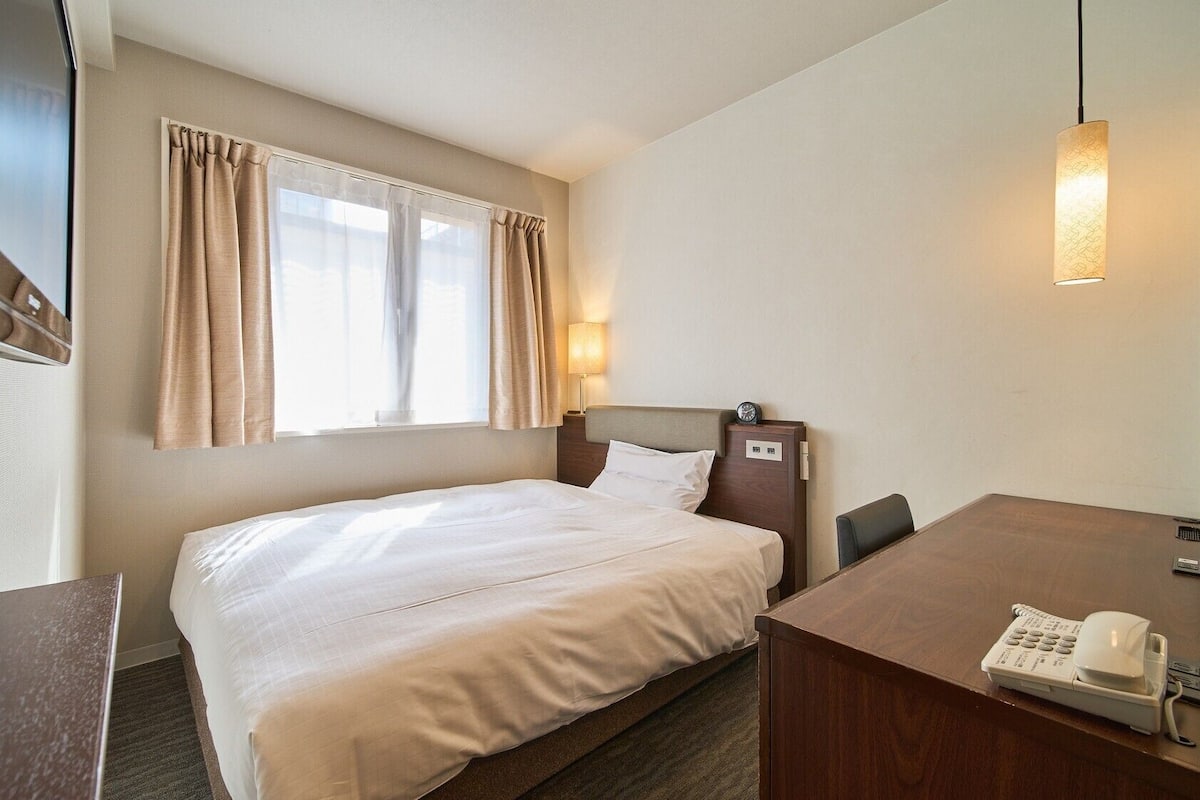 A hotel room with a bed, a desk with a telephone, a window with curtains, and a wall-mounted lamp.