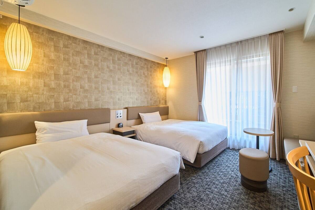 A hotel room with two beds, a desk, a chair, a TV, and a window with curtains.