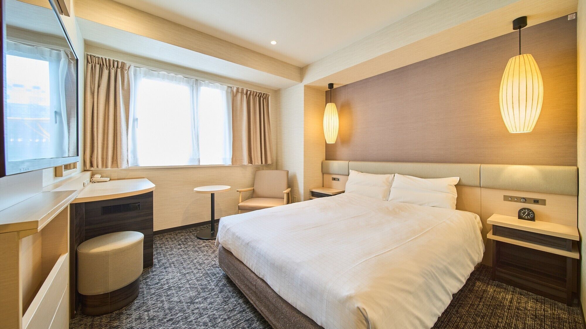 A hotel room with a large bed, a desk, a chair, a small table, and a bedside table with a clock.