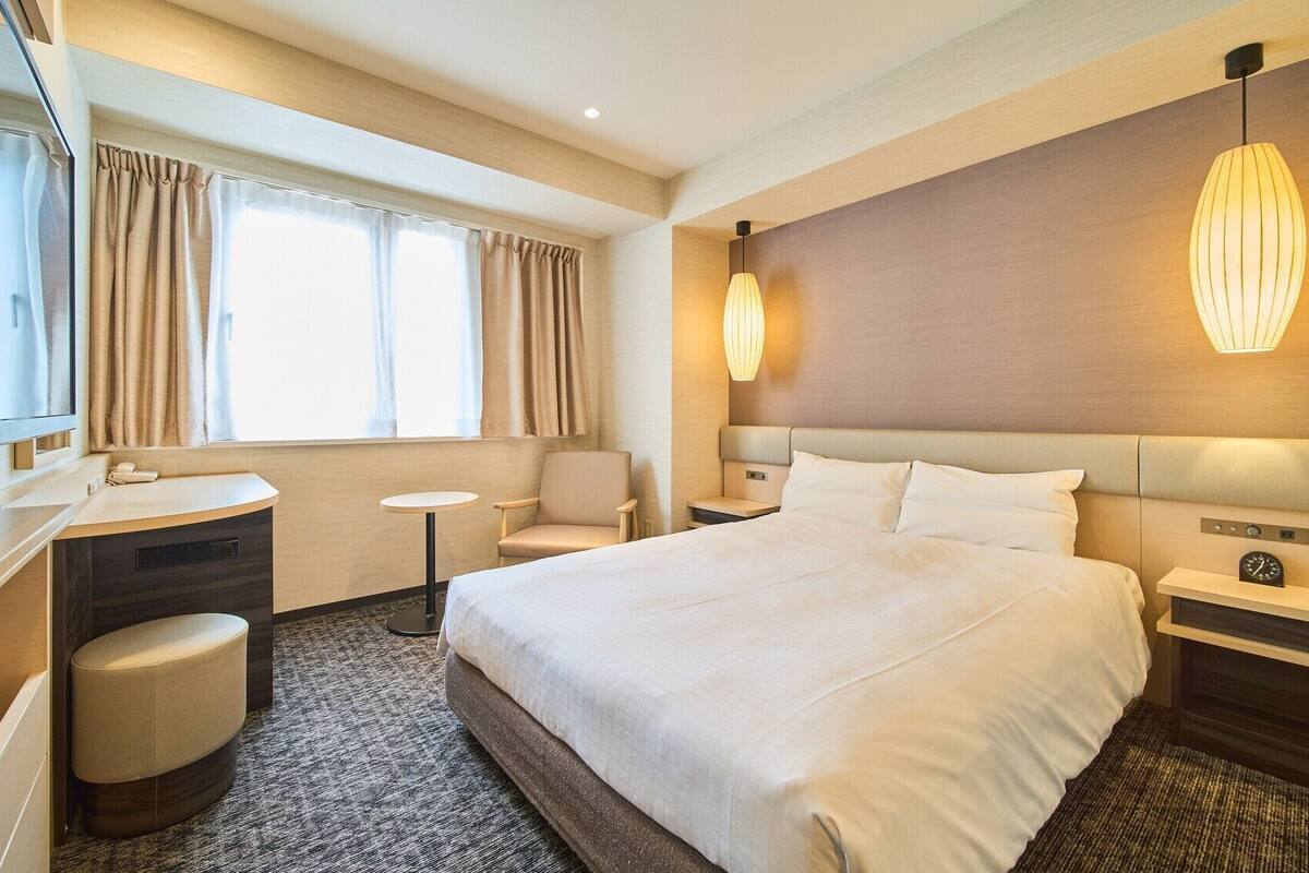A hotel room with a large bed, a desk, a chair, a small table, and a bedside table with a clock.