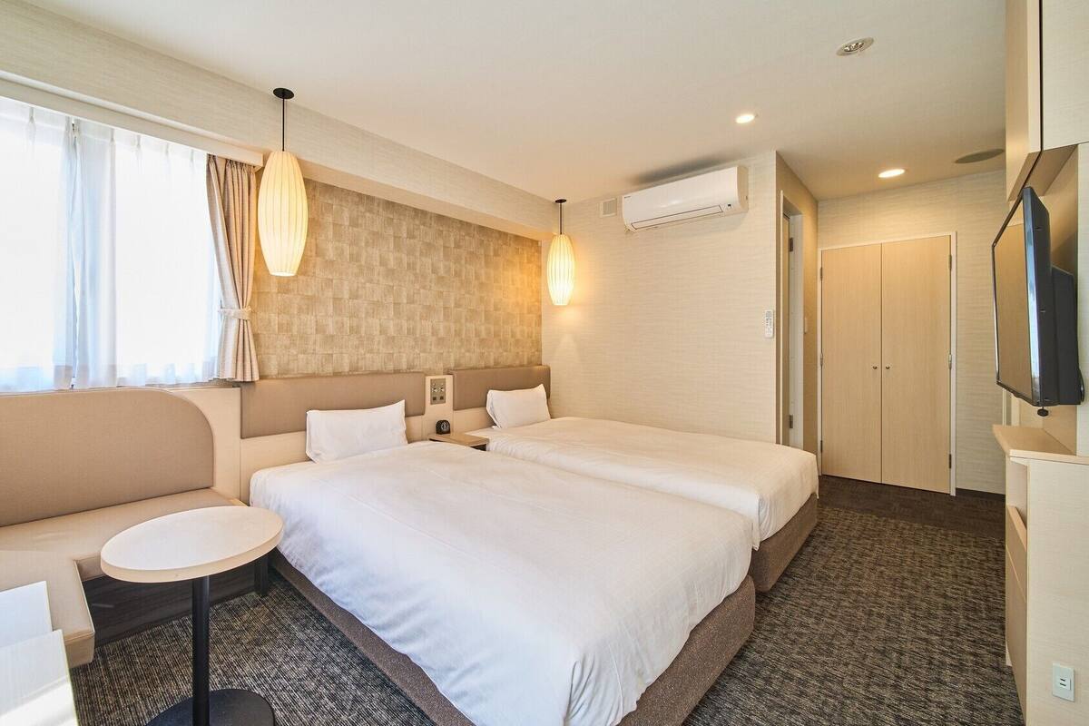 A hotel room with two beds, a sofa, a small table, a TV, and an air conditioning unit.