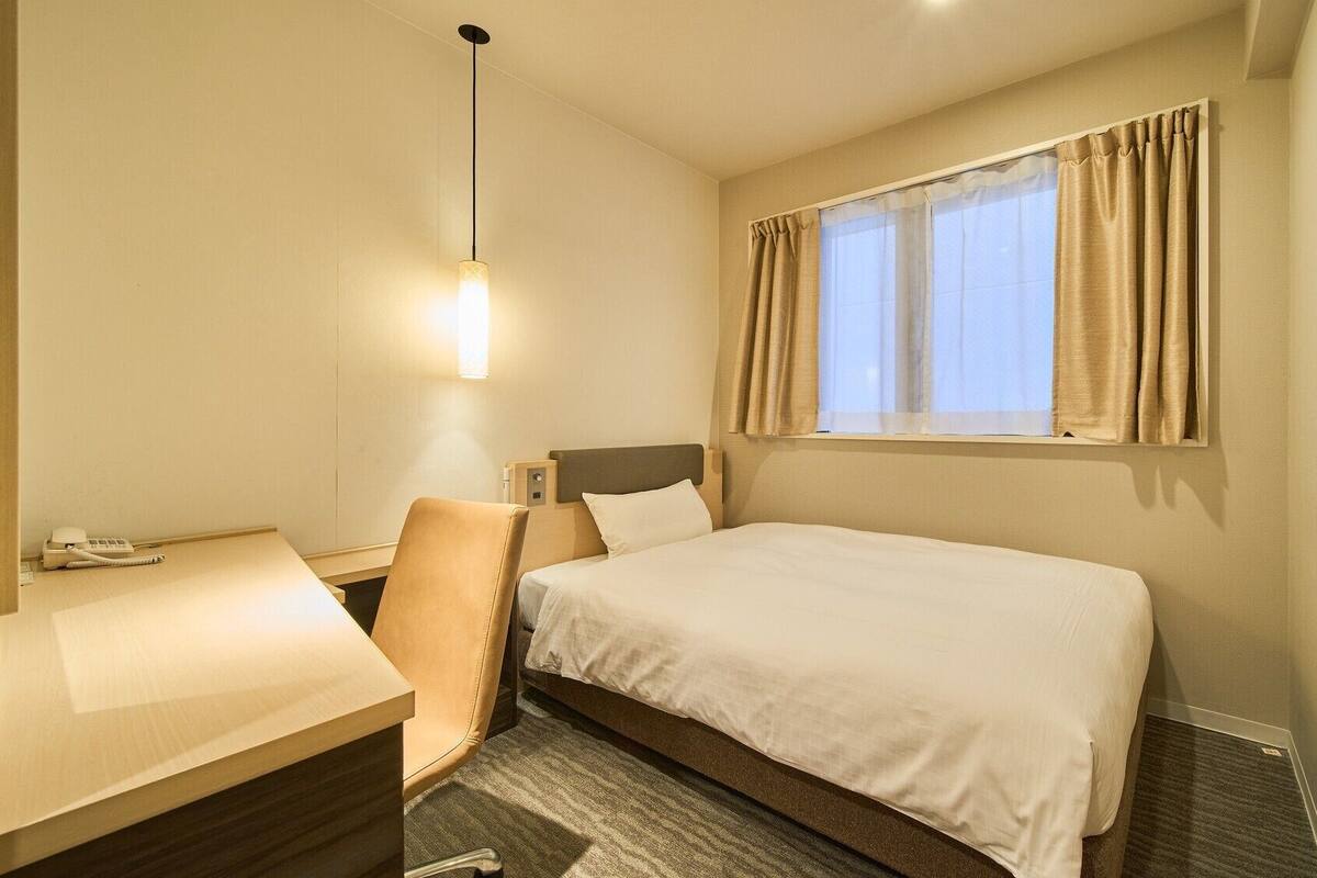 A hotel room with a bed, a desk, a chair, a telephone, a television, and a window with curtains.