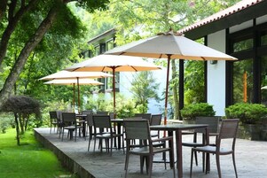 An outdoor patio with tables and chairs, surrounded by greenery and a building with large windows.