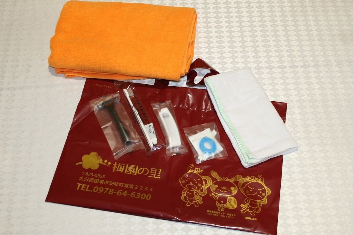 Una toalla naranja doblada con pulcritud, un bolso rojo con un emblema dorado y diversos artículos de aseo sobre una superficie estampada.