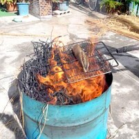 Un barril metálico con una parrilla en la parte superior, lleno de carbón encendido y un pescado asándose.