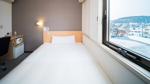 Habitación de hotel con cama, escritorio y silla, tetera y vistas a un paisaje urbano a través de una ventana.