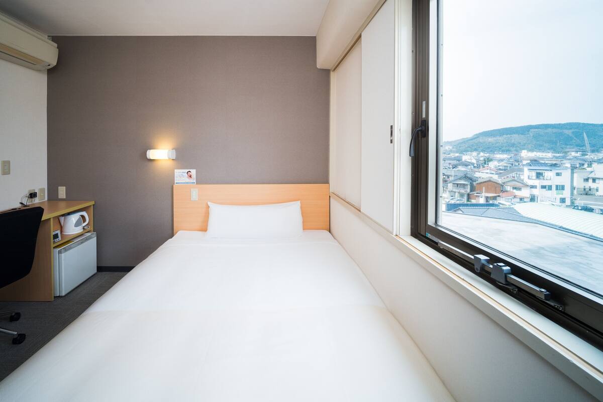 A hotel room with a bed, a desk with a chair, a kettle, and a view of a cityscape through a window.