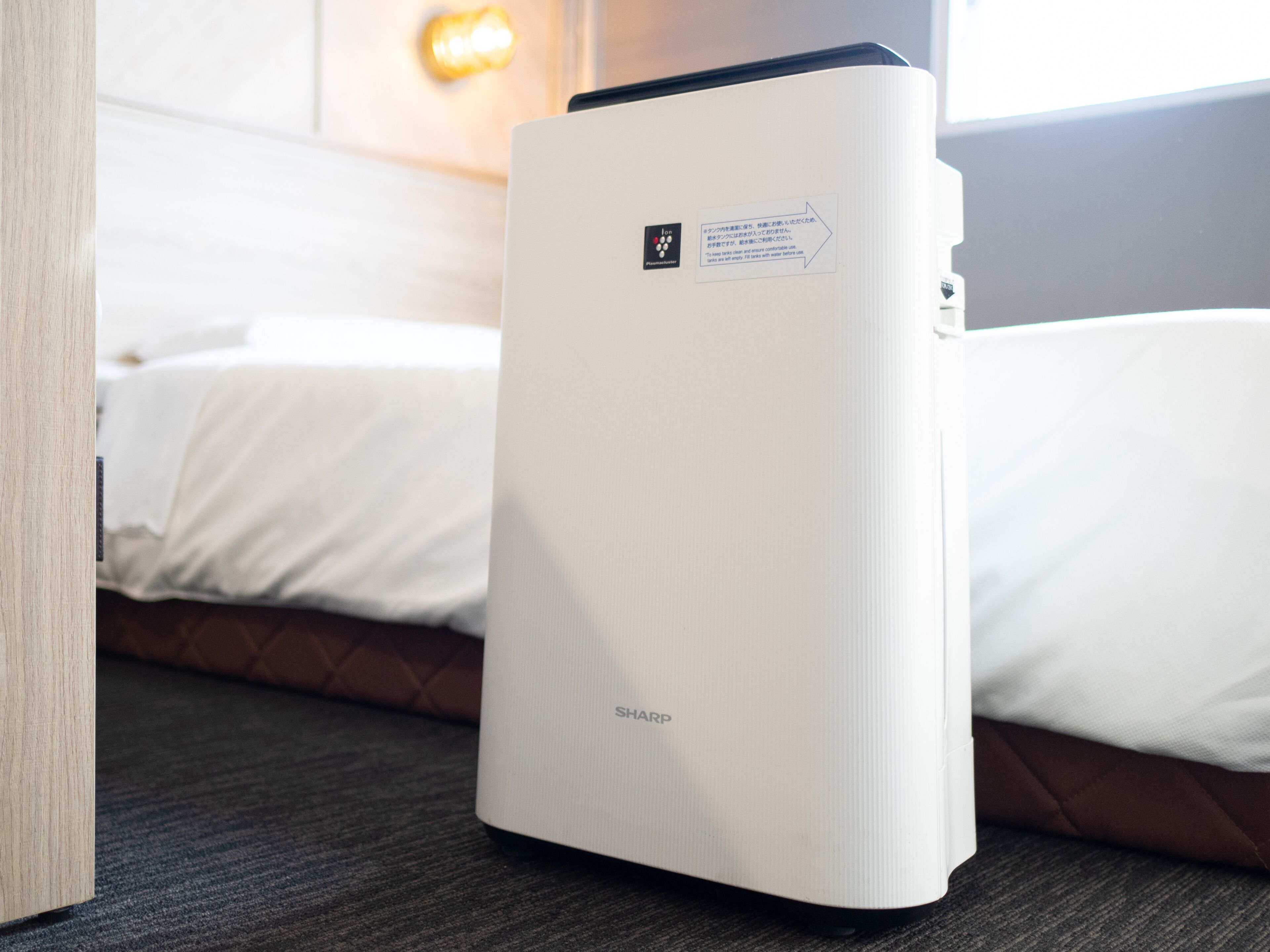 A white portable air conditioner in a hotel room with a bed and a wall-mounted light.
