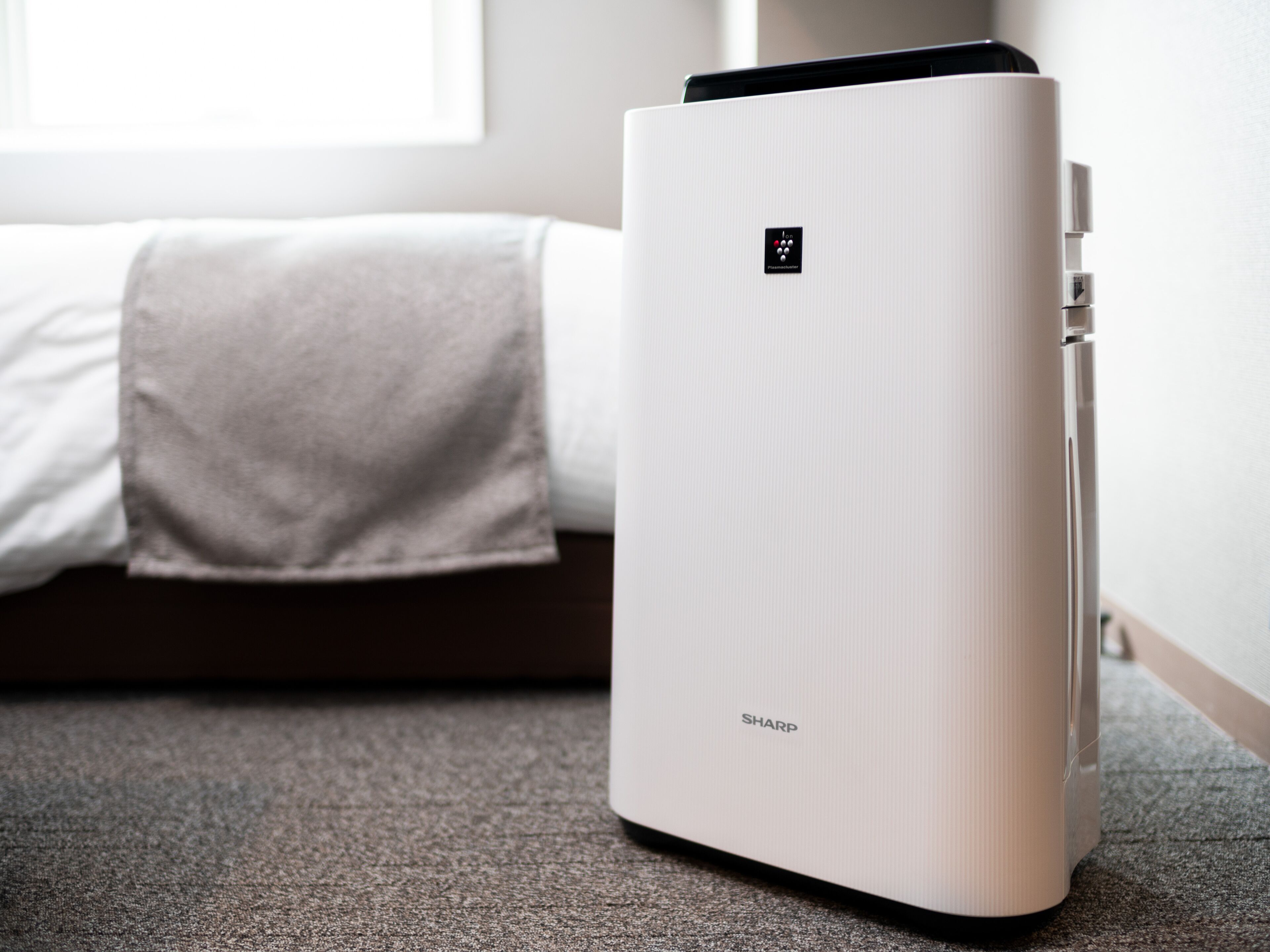 Um purificador de ar branco com painel de controle e a marca 'SHARP' em um chão coberto de carpete, ao lado de uma cama com cobertor branco.