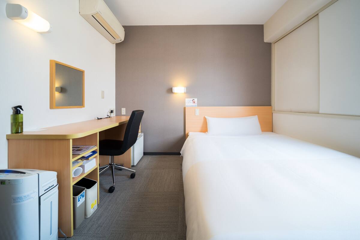 A hotel room with a bed, a desk with a chair, a mirror, and an air conditioning unit.