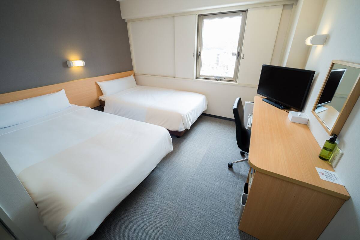 A hotel room with two beds, a desk with a television, a chair, a window, and a wall-mounted light.