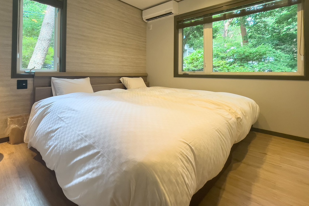 A bedroom with a large bed, two windows, a bedside table, and a wall-mounted air conditioner.