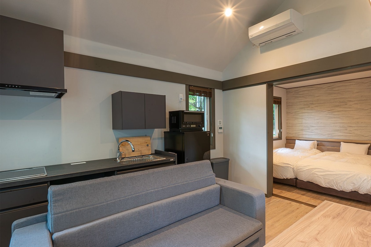A modern, compact bedroom with a large bed, a small desk, and a grey sofa.