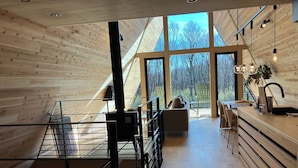 Un interior moderno con piso de madera, una zona de cocina, un rincón de descanso con sofá y ventanas amplias que ofrecen vistas a árboles.