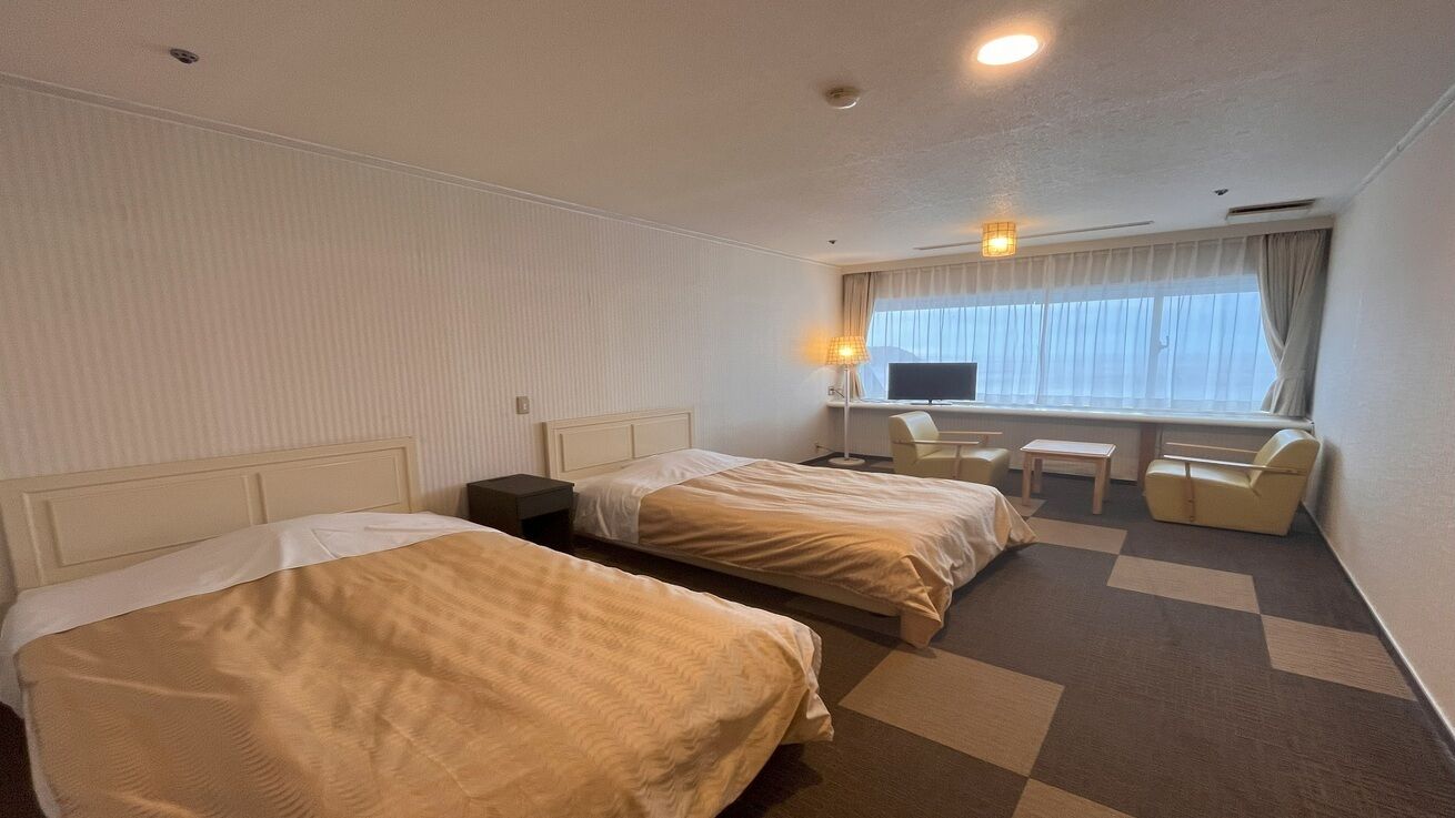 Ein Hotelzimmer mit zwei Betten, einem Schreibtisch, einem Stuhl, einem kleinen Tisch und einem Fenster mit Vorhängen.