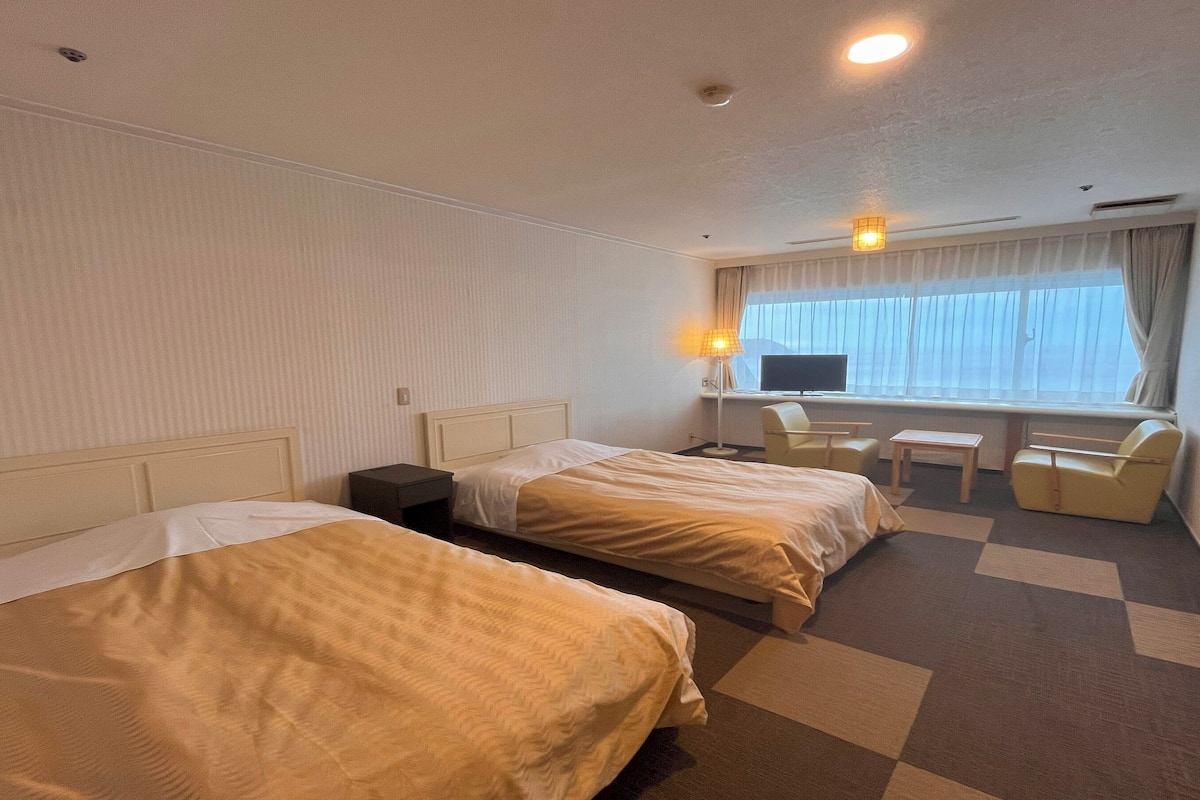 Quarto de hotel com duas camas, uma escrivaninha, uma cadeira, televisão e janela com cortinas.
