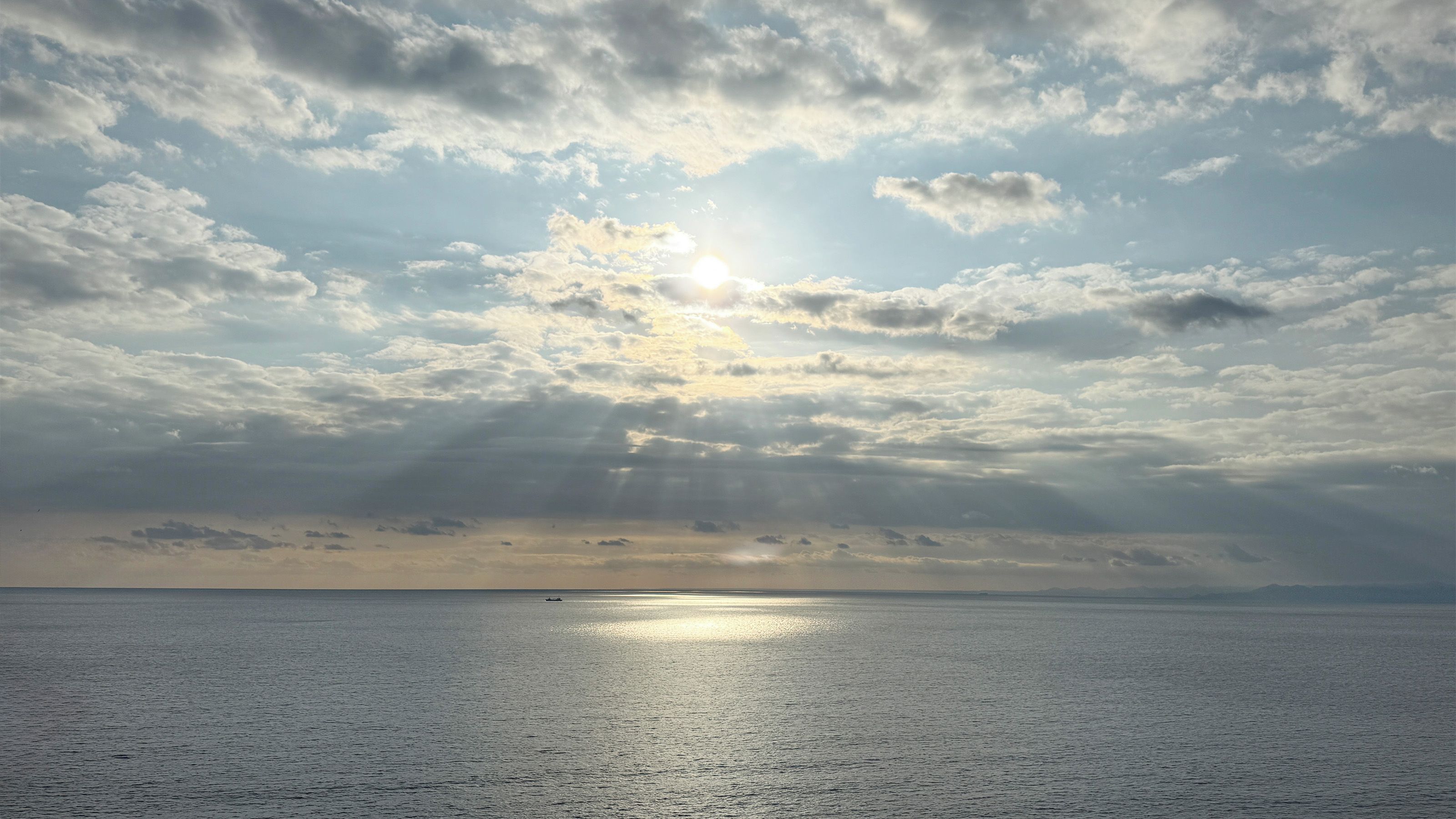 A calm sea with sunlight breaking through clouds, a distant horizon, and a small boat.