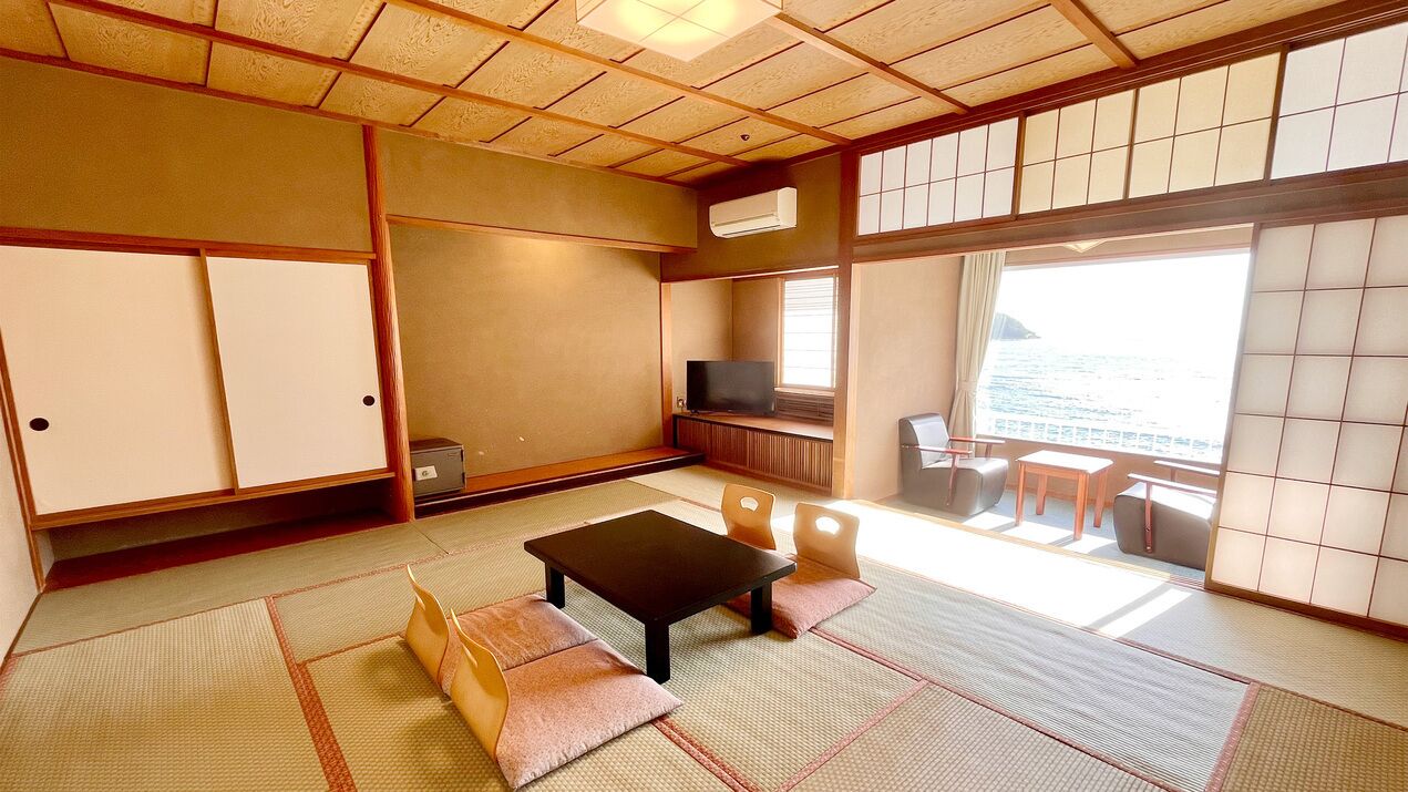 Ein traditioneller japanischer Raum mit Tatami-Boden, einem niedrigen Tisch, Stühlen, einem Fernseher und einer Schiebetür zu einem Balkon mit Aussicht.