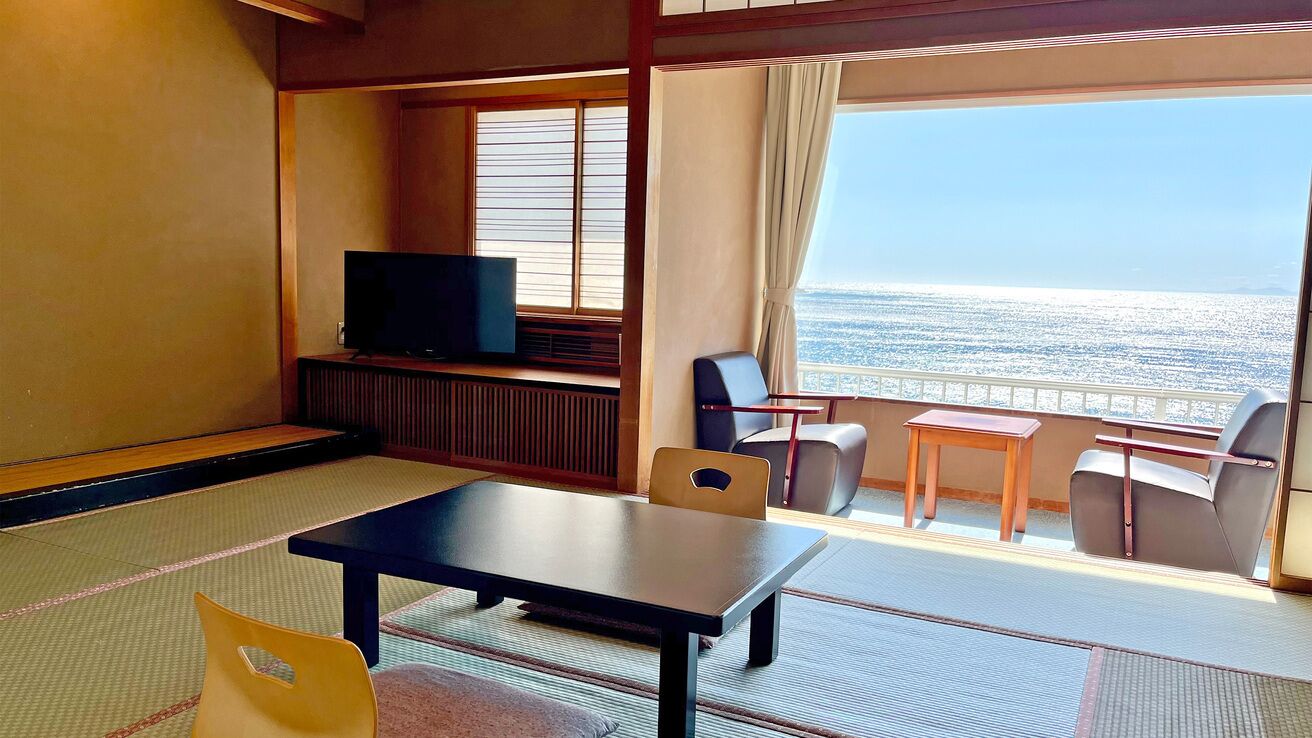 Ein Zimmer mit Tatami-Böden, einem Fernseher, einem Tisch, Stühlen und Meerblick.