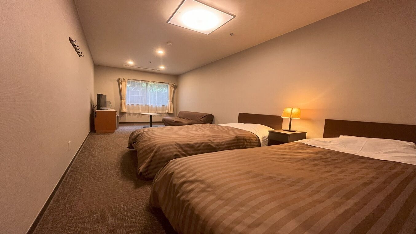 Ein Hotelzimmer mit zwei Betten, einem Schreibtisch, einem Stuhl, einer Lampe und einem Fenster mit Vorhängen.