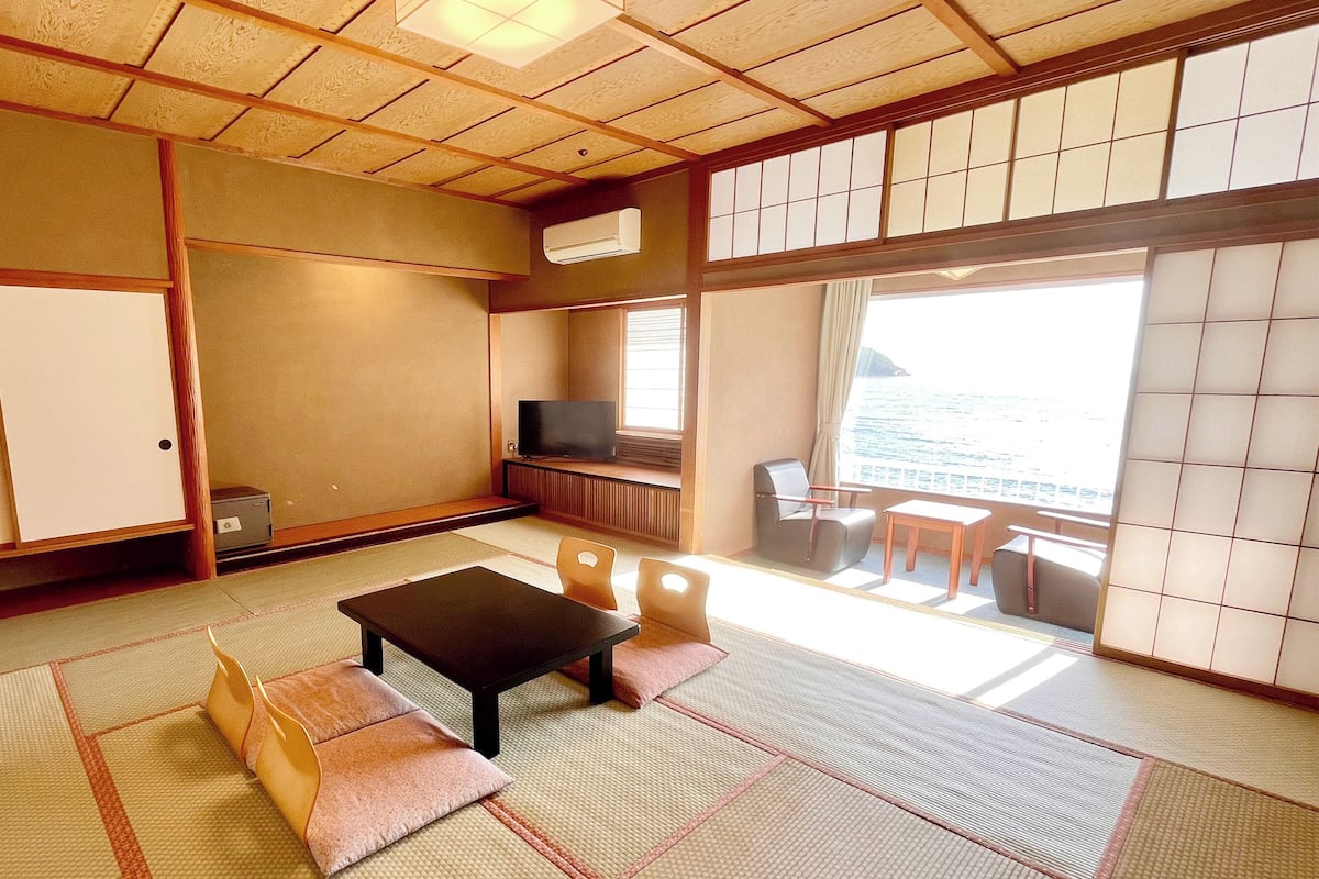 Um quarto japonês tradicional com tatames, uma mesa baixa, cadeiras, uma televisão e vista para o mar.