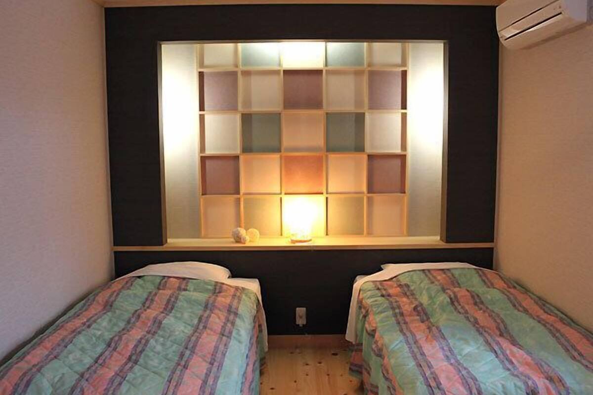 A room with two beds, a window with a light, and a wall-mounted air conditioner.